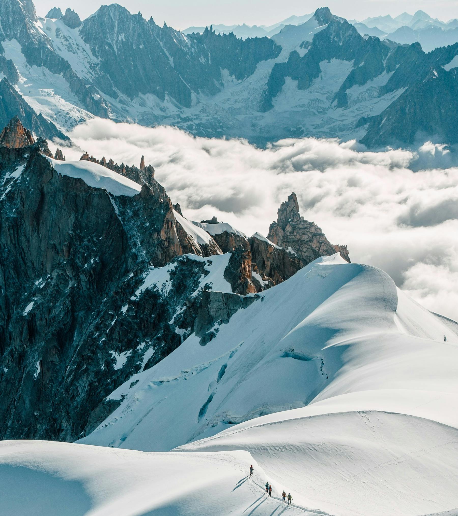 Cordées progressant en file sur une arête de neige au-dessus d'une mer de nuages, face aux aiguilles du massif du Mont-Blanc