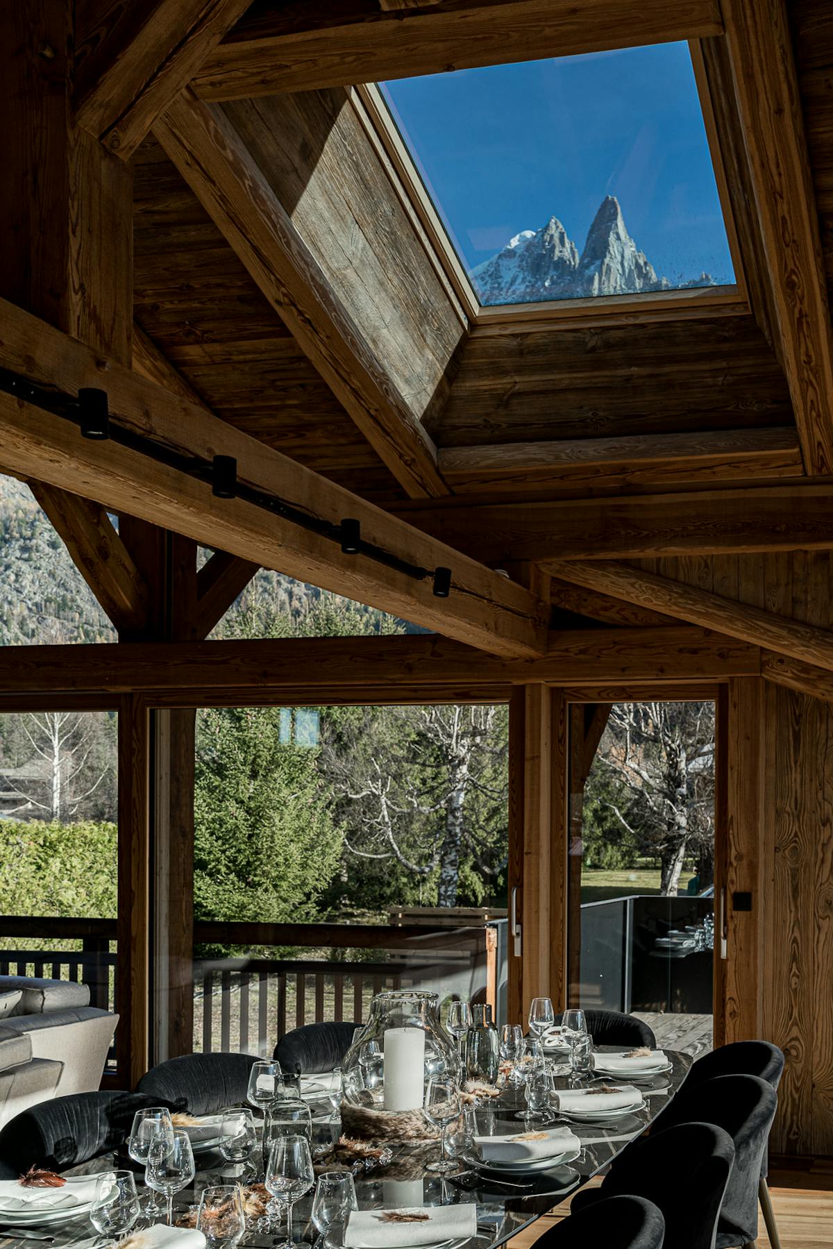 The big table at Chalet Bonami laid under the old-timber roof frame, the roof window open on the Drus, and beyond the glazed wall the balcony table