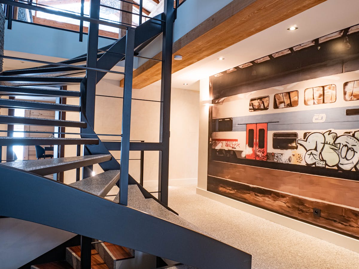 Straight black metal staircase with stone treads linking the levels of a chalet, in front of a wall mural of a graffitied train carriage