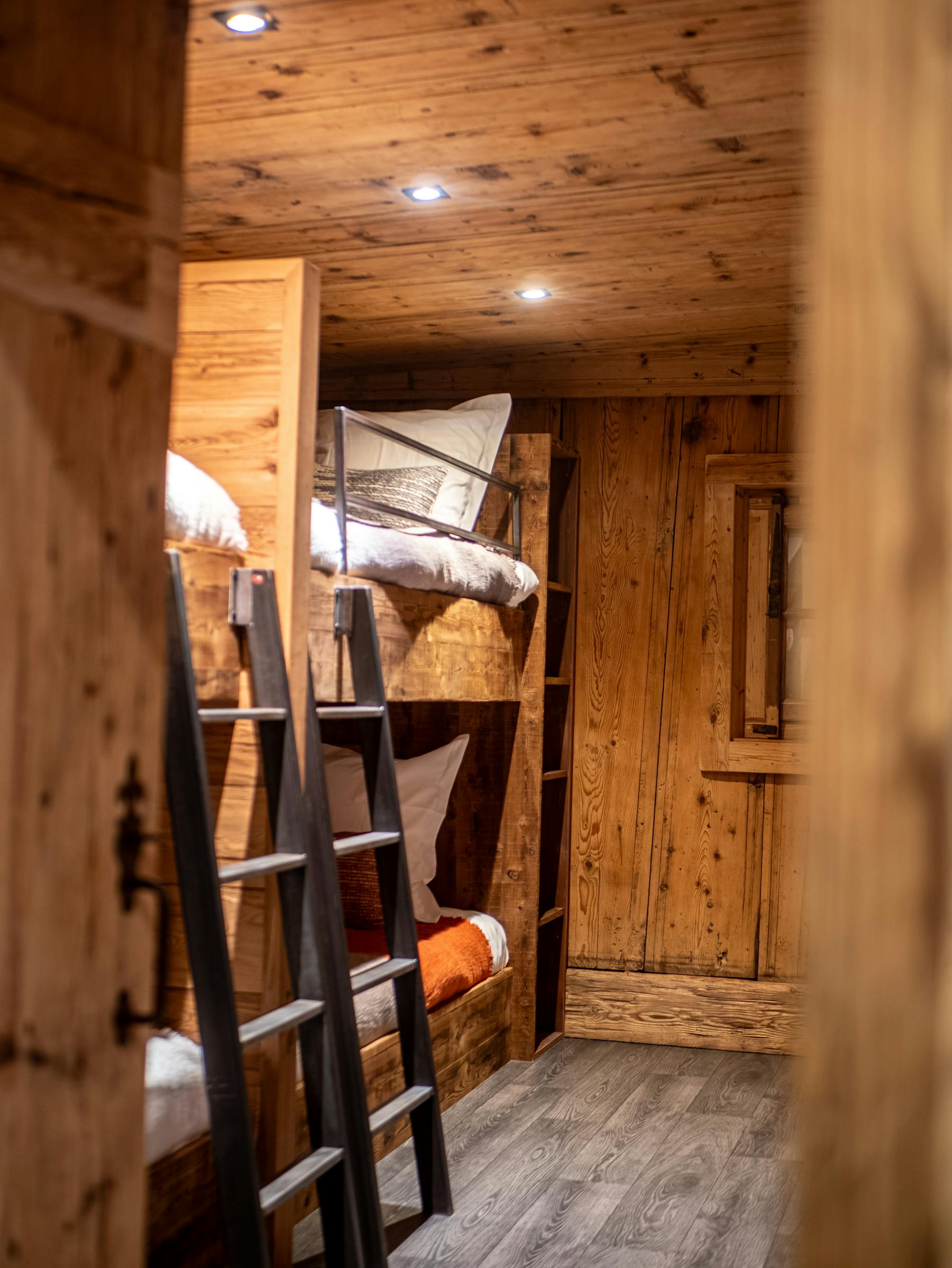 Two bunk beds in old timber with black metal ladders, in the bunk room of a chalet, seen through a half-open door