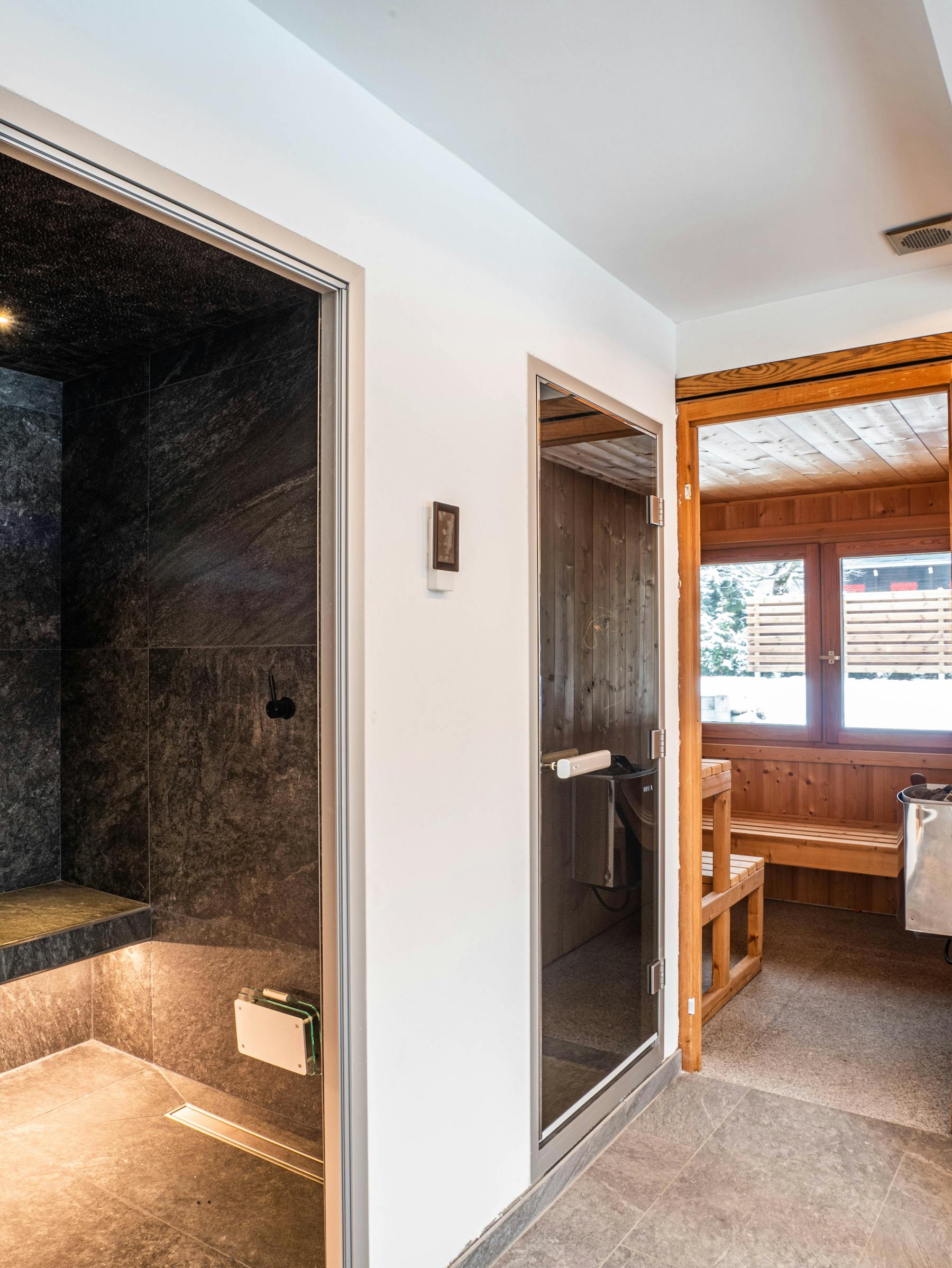 Le spa du Chalet Bonami aux Bois : l'entrée vitrée du hammam en pierre sombre, la cabine de douche, et le sauna traditionnel en bois dont la fenêtre donne sur la neige
